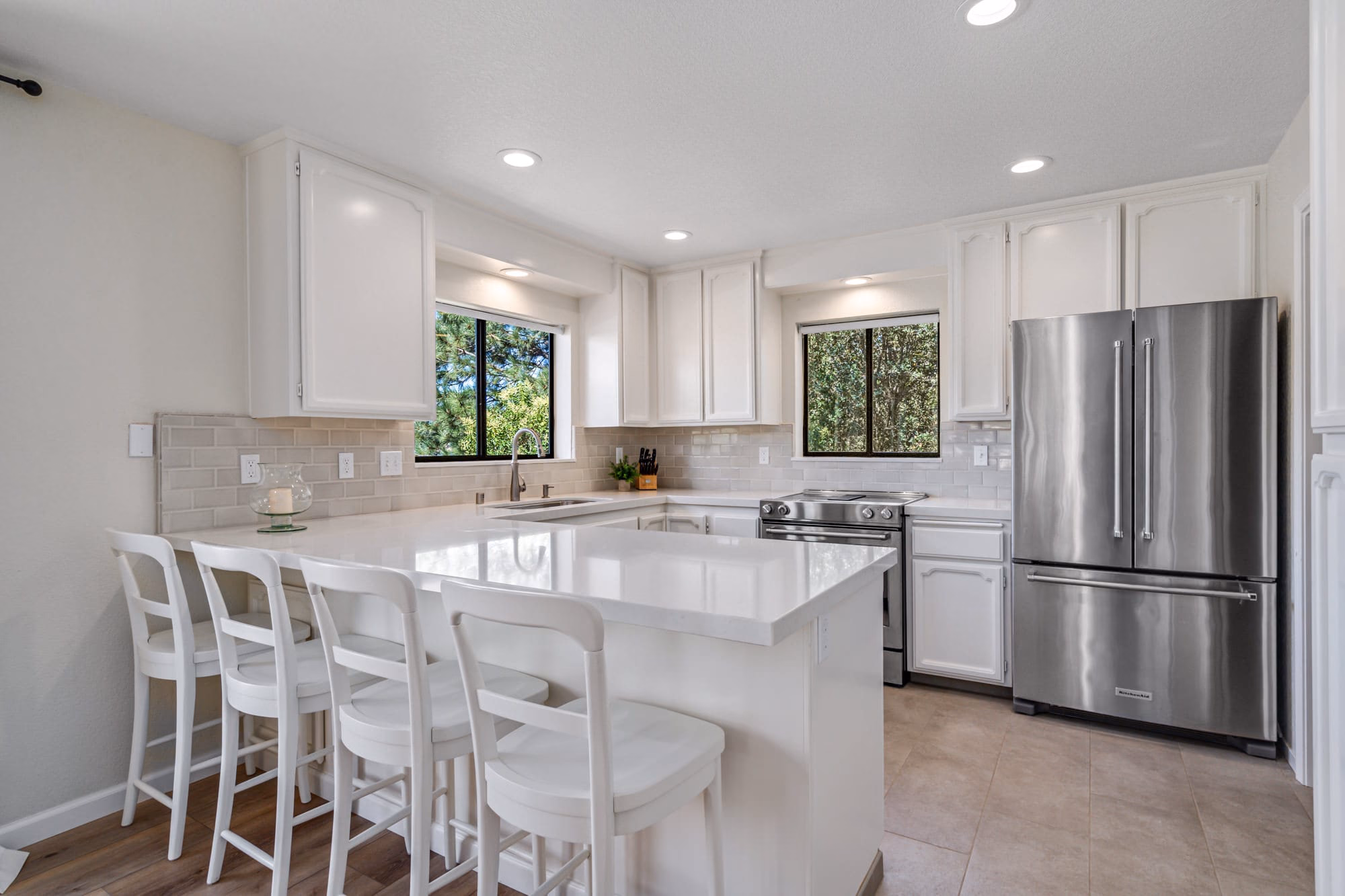 White kitchen with a large breakfast bar, four stools, stainless-steel appliances and windows facing trees