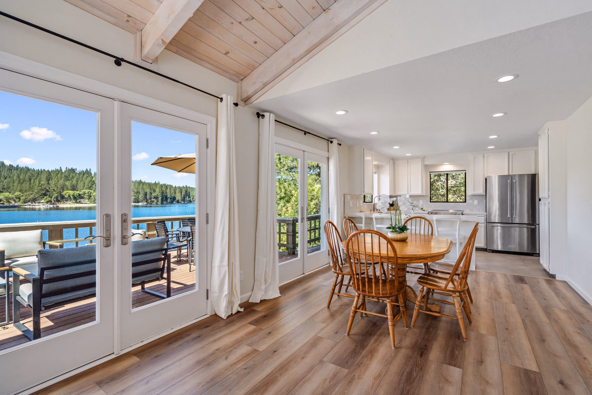 Open dining room and white kitchen beside French doors leading to a furnished lakefront deck