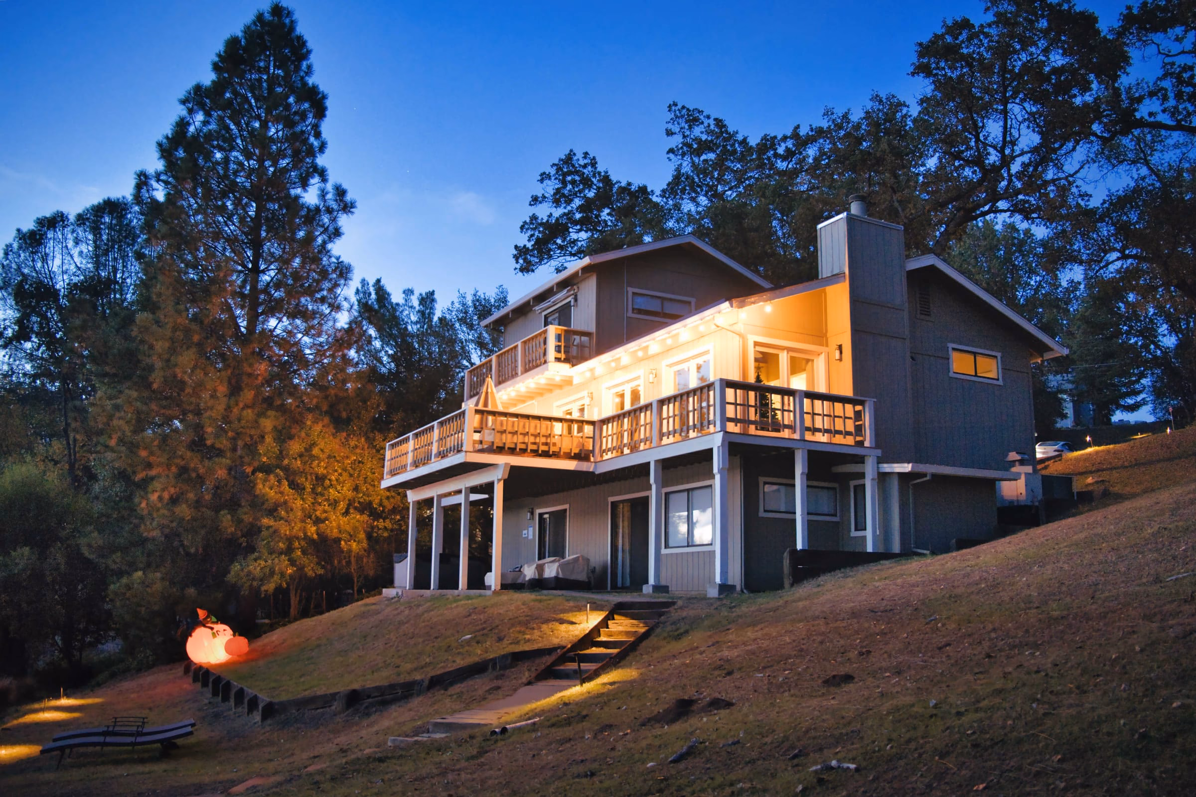 Three-storey lakefront house with its decks lit at dusk among tall pine and oak trees
