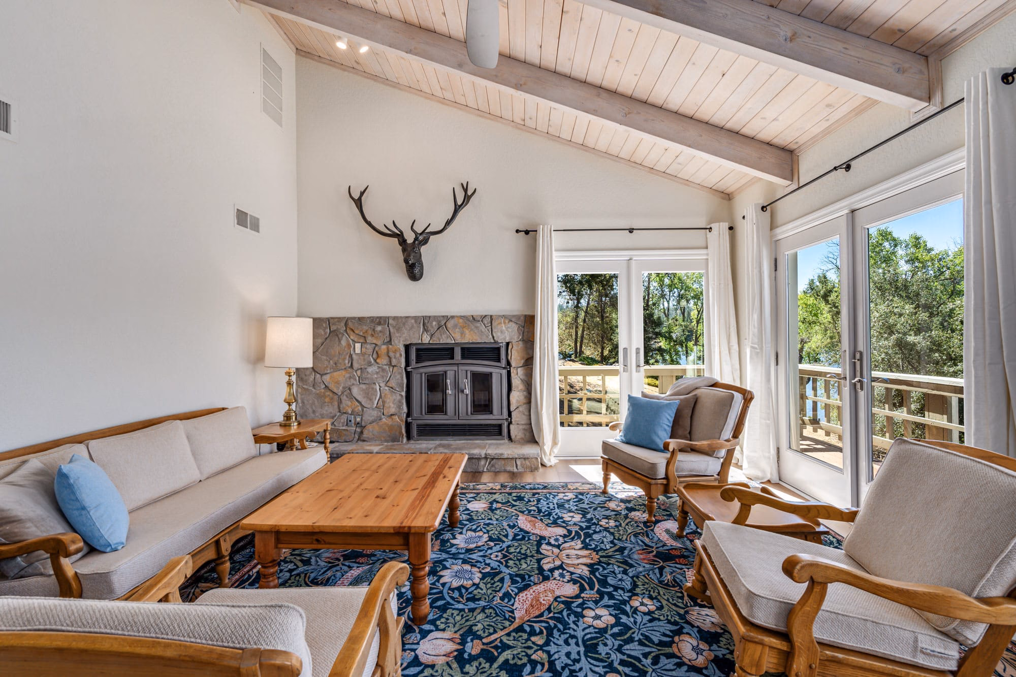 Bright living room with a stone fireplace, vaulted wood ceiling, sofas and French doors opening toward the lake